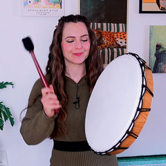 A woman with long brown hair holds a hand drum with a white drumhead and a wooden rim, while holding a drumstick in a home setting with framed art on the wall.
